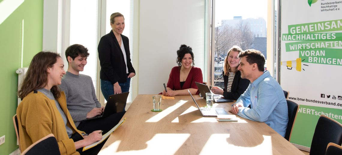 Mitarbeiter:innen des BNW sitzen an einem Konferenztisch und besprechen sich. Durch die offenen Fenster scheint die Sonne im Hintergrund steht ein BNW-Rollup