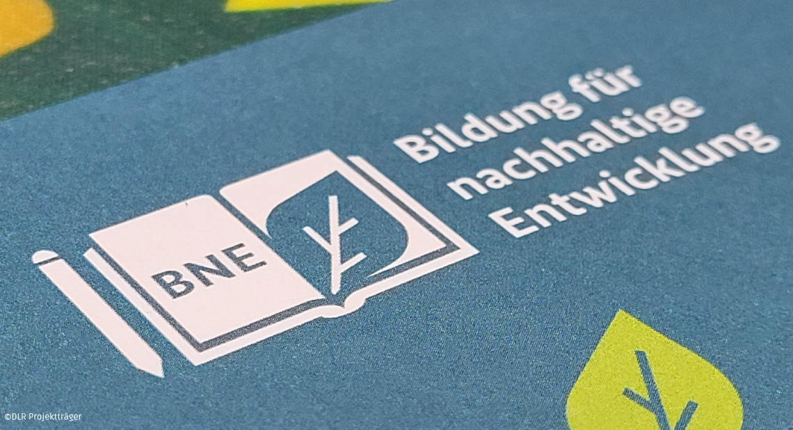 BNE-Logo: Aufgeschlagenes Buch mit ‚BNE' auf der linken Seite und einem Blatt-Symbol auf der rechten Seite. Text: ‚Bildung für nachhaltige Entwicklung'. Blauer Hintergrund mit gelben und grünen Blatt-Elementen. Quelle: DLR Projektträger.