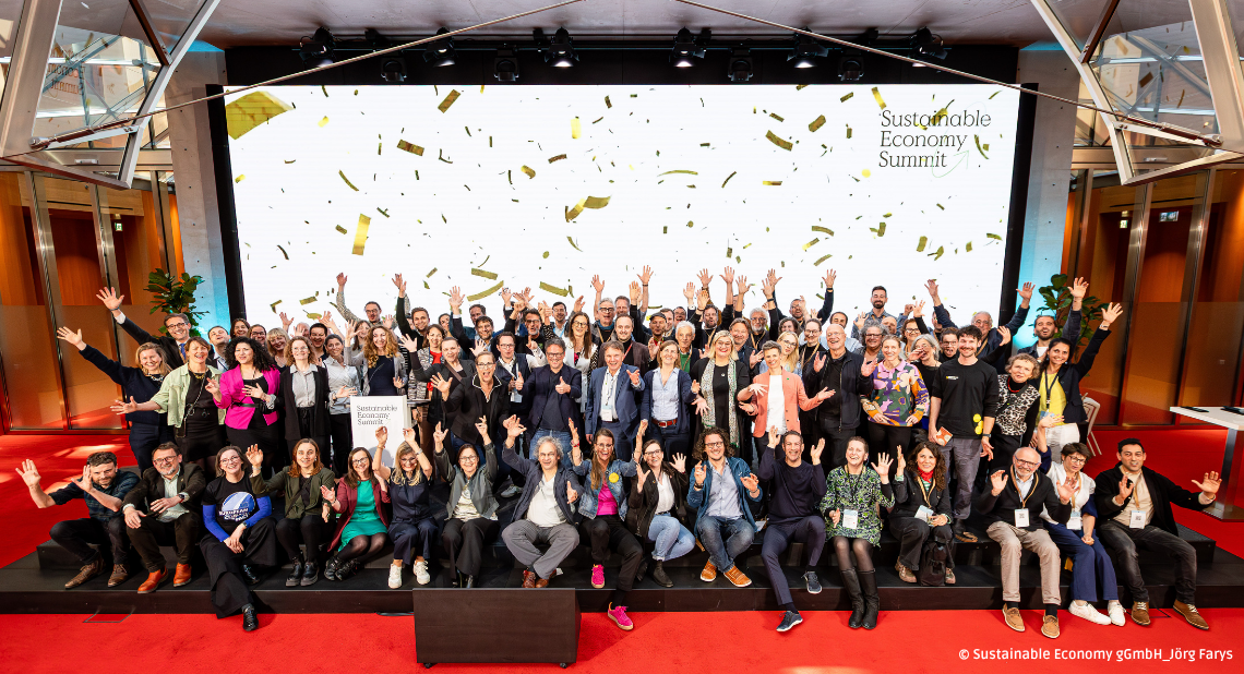 Sustainable Economy Summit 2026: Etwa 100 Teilnehmer in Geschäftskleidung stehen auf einer roten Bühne vor einer großen Leinwand, viele mit erhobenen Händen und Armen. Goldene Konfetti fällt von oben.