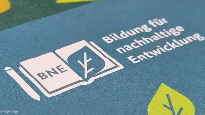 BNE-Logo: Aufgeschlagenes Buch mit ‚BNE' auf der linken Seite und einem Blatt-Symbol auf der rechten Seite. Text: ‚Bildung für nachhaltige Entwicklung'. Blauer Hintergrund mit gelben und grünen Blatt-Elementen. Quelle: DLR Projektträger.