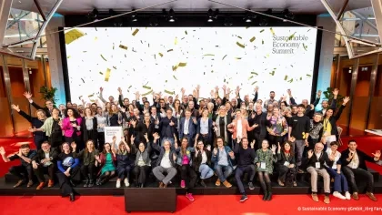 Sustainable Economy Summit 2026: Etwa 100 Teilnehmer in Geschäftskleidung stehen auf einer roten Bühne vor einer großen Leinwand, viele mit erhobenen Händen und Armen. Goldene Konfetti fällt von oben.