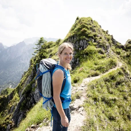 Antje von Dewitz auf einem Wanderpfad in den Bergen mit einem Rucksack mit Kletterseilen auf dem Rücken lächelt in die Kamera